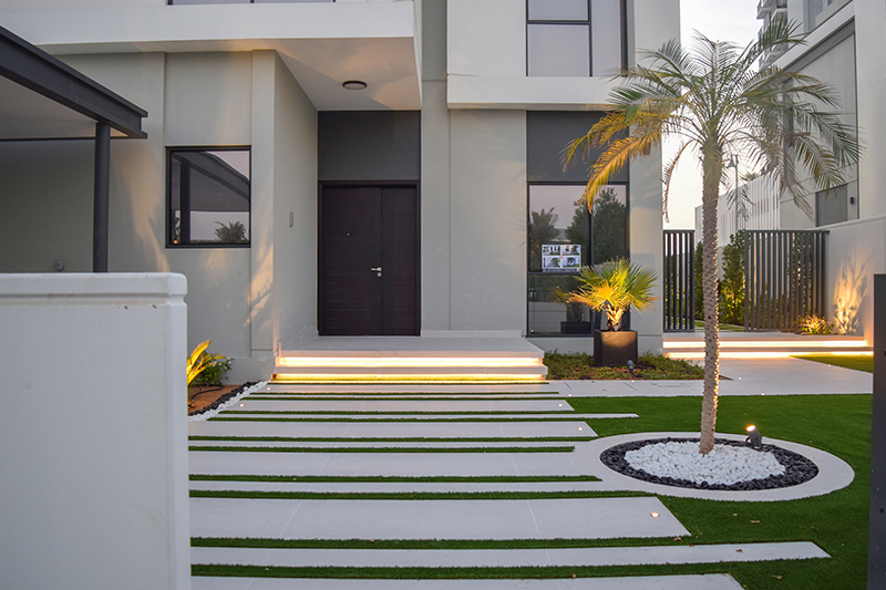 Palm tree in circular white stone bed, lit concrete entrance steps, grass and concrete path, and dark wooden door in a contemporary Dubai landscape
