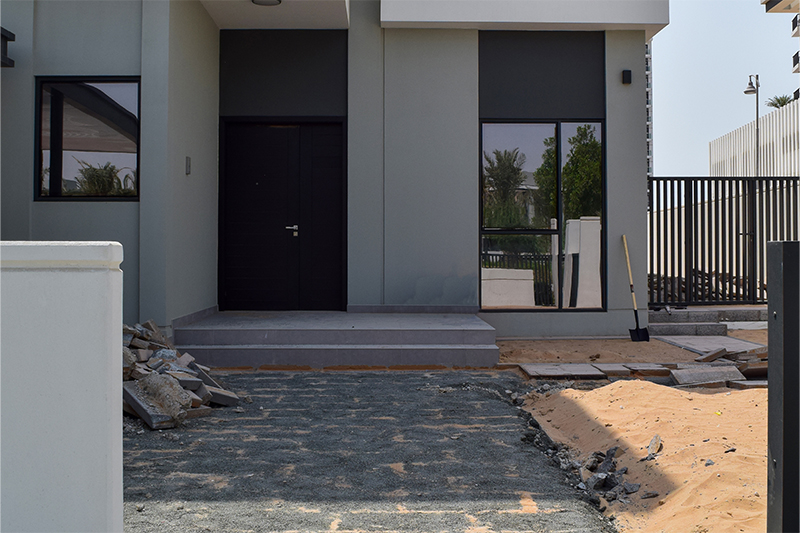 Under-construction modern home entrance with dark front door, large windows, and landscape materials on-site in Dubai.