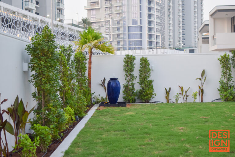 Elegant landscaped garden with lush plants, tall palm tree, sculptural blue vase, and manicured lawn set against modern high-rise buildings in Dubai.