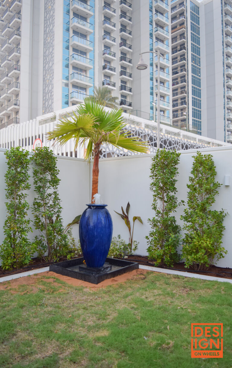 Tall blue ceramic water fountain surrounded by lush plants, tropical greenery, and neatly trimmed lawn in a modern Dubai garden.