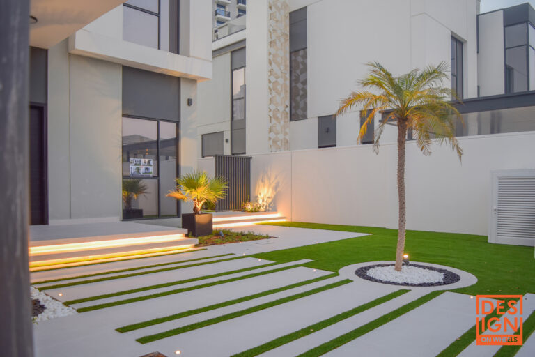 Modern minimalist garden with grass-inlaid pathways, palm trees, ambient lighting, and structured tropical landscaping by Design on Wheels.