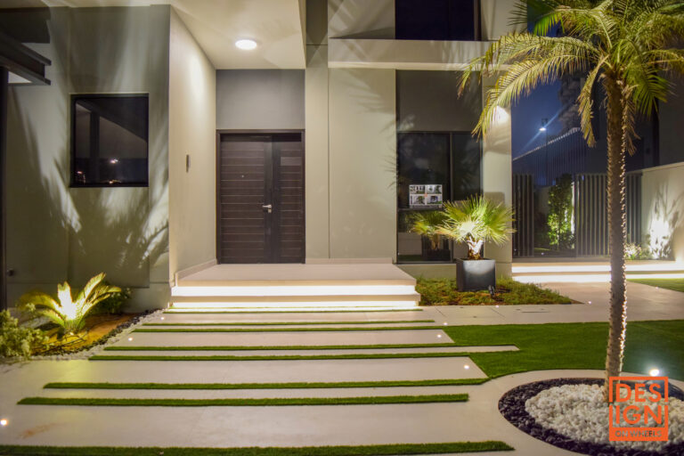 Contemporary house entrance with geometric grass-and-concrete path, illuminated steps, and potted tropical plants by Design on Wheels.