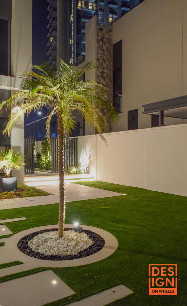 Palm tree in modern garden illuminated at night, surrounded by white and black stones, artificial turf, and contemporary architecture. Caption: Dramatic night-time garden design in Dubai with palm centerpiece, layered stone textures, and soft landscape lighting—executed by Design on Wheels. Description: This image features a premium modern garden design executed by Design on Wheels in Dubai. At its center, a majestic palm tree rises from a sculptural circular bed of black and white decorative stones, lit dramatically for nighttime ambiance. Surrounding the feature is an expanse of manicured artificial grass, framed by clean architectural lines and a gated entrance. This project integrates nature, geometry, and lighting to craft a serene and visually striking outdoor space—ideal for showcasing high-end landscape execution, residential garden design, and outdoor lighting innovation across the UAE. Want help linking this content to a services page, writing Google snippets, or building out an SEO-rich landing section? I’ve got just the toolkit.