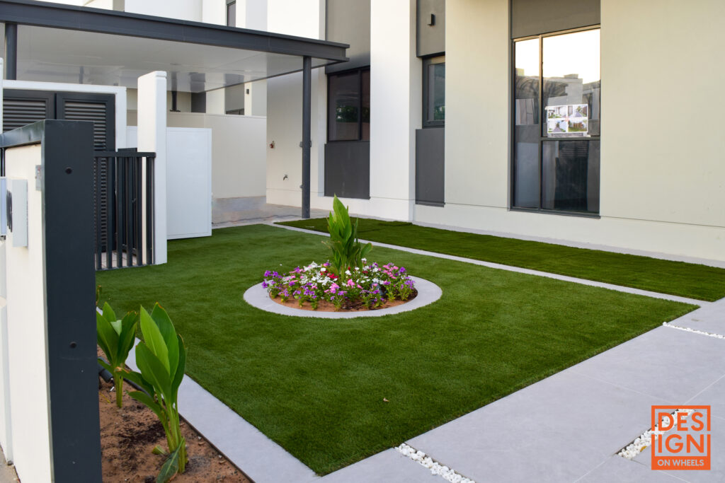 Contemporary garden with artificial grass, circular flower bed, tropical plants, and a modern glass-walled structure by Design on Wheels.
