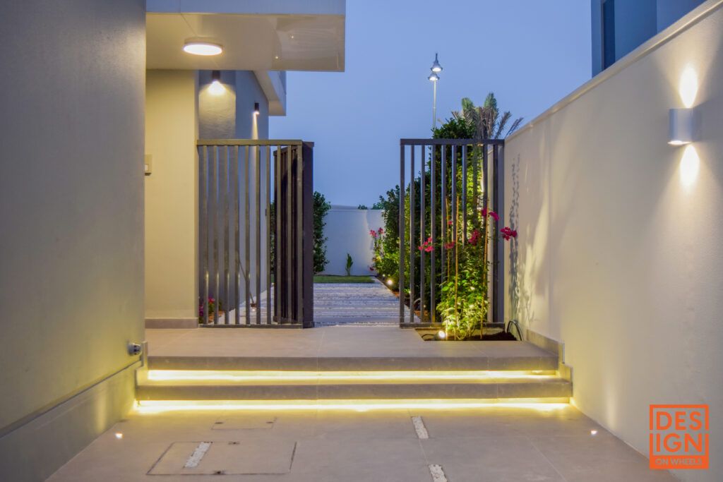 Illuminated modern garden entrance with step lighting, sleek gate, lush greenery, and floral borders by Design on Wheels. Caption: Stylish landscape design in Dubai showcasing glowing entrance steps, tropical planting, and a contemporary gate—executed by Design on Wheels. Description: This image features a sophisticated landscape execution in Dubai by Design on Wheels, highlighting a well-lit garden entrance with ambient step lighting and sleek architectural lines. The design includes structured planting with lush greenery, vibrant flowers, and modern outdoor lighting that creates a warm, welcoming atmosphere. A clean concrete path leads to a contemporary black gate, framed by vertical lighting and manicured tropical borders. This project demonstrates the perfect blend of aesthetics and functionality—ideal content for websites focused on villa landscaping, outdoor lighting solutions, and premium garden design in the UAE and GCC region. Would you like me to build this into a showcase layout or optimize it for social platforms like Pinterest or Instagram too?