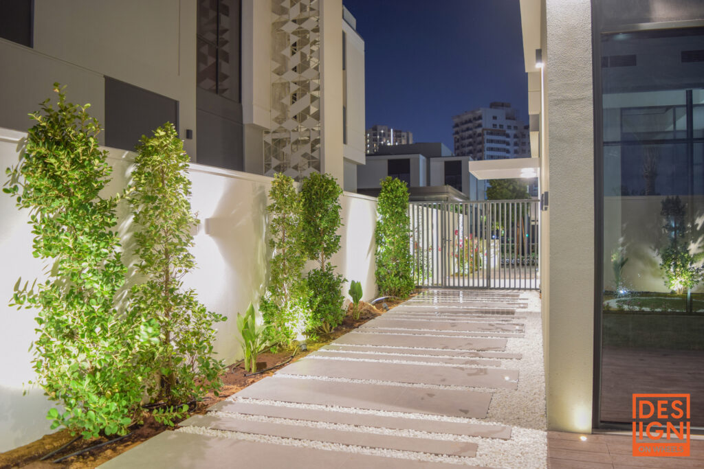 Contemporary garden path with geometric tiles, white gravel, lush green plants, and soft ambient lighting in a modern residential setting.