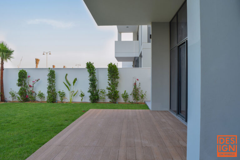 Modern backyard with wooden deck, green lawn, white privacy wall, and structured tropical landscaping by Design on Wheels.