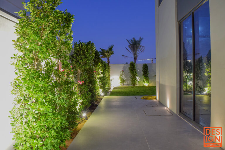 Elegant side garden of a modern villa in Dubai with layered plants, landscape lighting, and a tiled walkway.